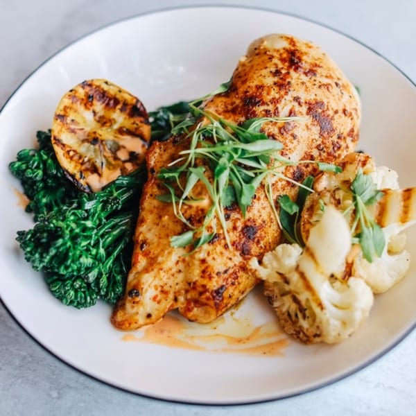 Grilled chicken breast with broccolini, roasted cauliflower, and a grilled lemon on a white plate.