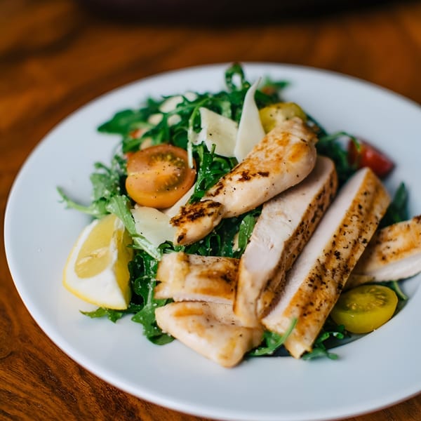 Grilled chicken salad with arugula, cherry tomatoes, parmesan, and lemon on a white plate.
