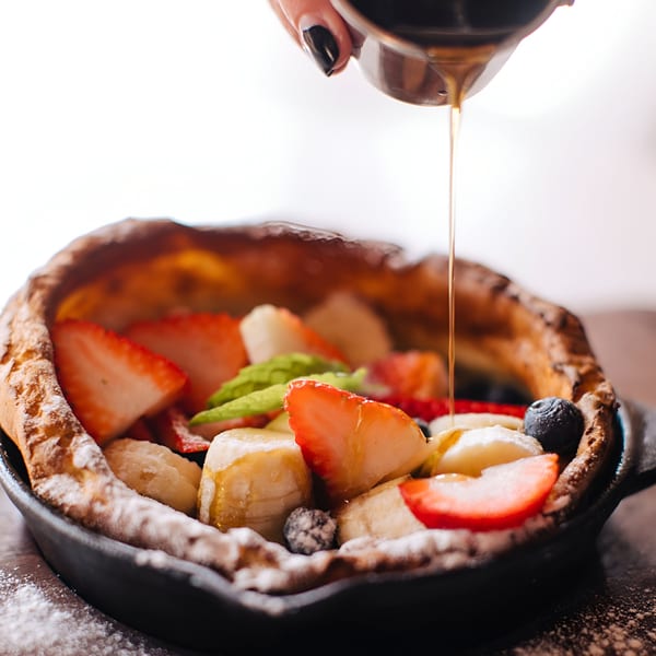 Syrup pouring over a Banyan Kitchen Dutch baby pancake with strawberries, bananas, and blueberries.