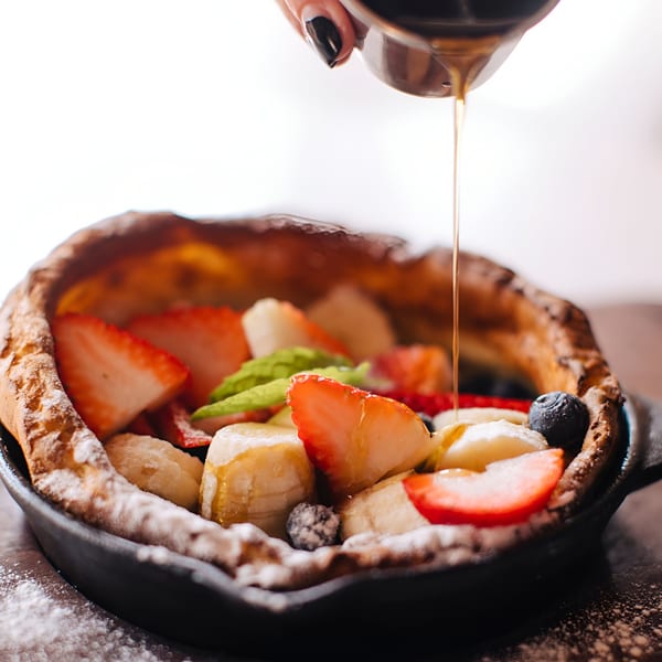 Maple syrup pouring over a Dutch baby pancake with strawberries, bananas, and blueberries.
