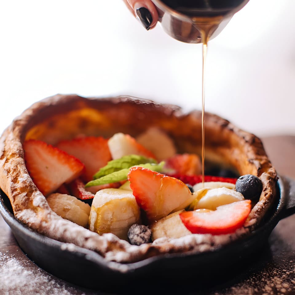 Maple syrup pouring over a Dutch baby pancake with strawberries, bananas, and blueberries.