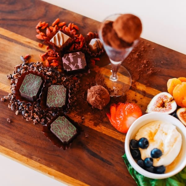 A wooden board with chocolates, goji berries, cocoa nibs, strawberries, figs, and a dessert bowl.