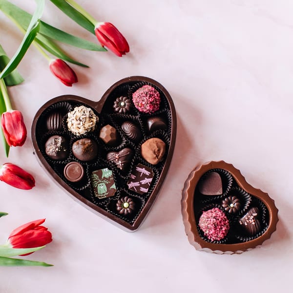 Two heart-shaped boxes of assorted chocolates and red tulips on a light pink surface.