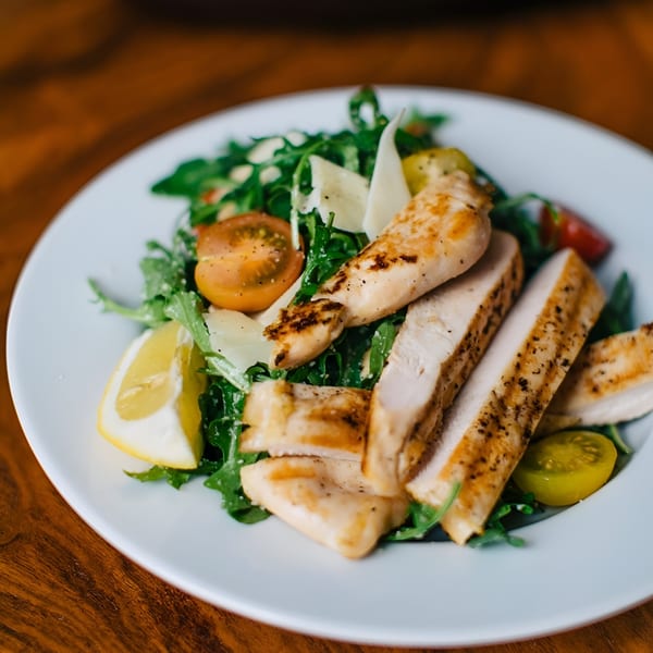 Grilled chicken salad with arugula, cherry tomatoes, Parmesan shavings, and a lemon wedge.