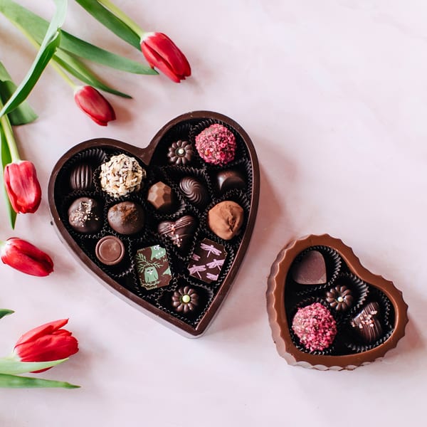 Two heart-shaped boxes of assorted chocolates and red tulips on a light pink surface.