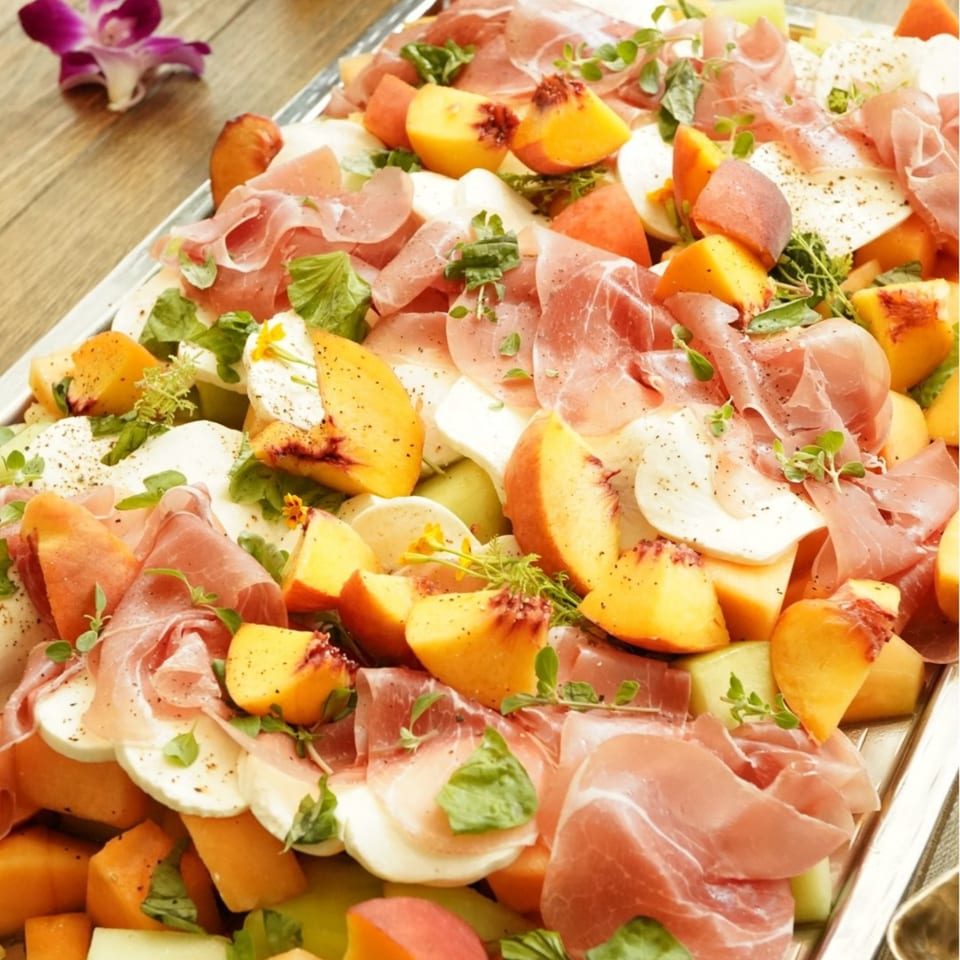 A platter of prosciutto, mozzarella, peaches, and melon, garnished with herbs and pepper.
