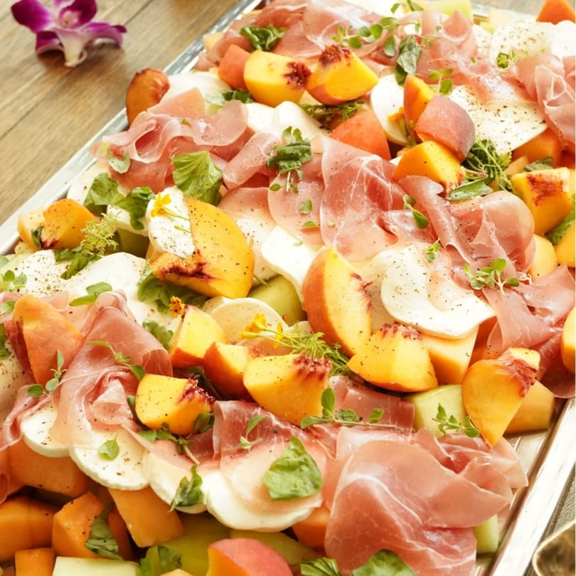 A platter of prosciutto, mozzarella, peaches, and melon, garnished with herbs and pepper.